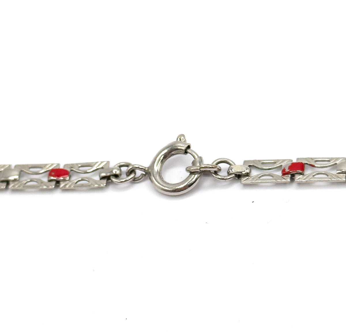 Close-up on the round spring ring clasp and its jump ring, bridging two runs of pierced flat links, each with a small red enamel square.