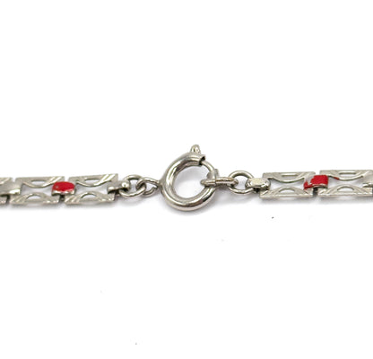 Close-up on the round spring ring clasp and its jump ring, bridging two runs of pierced flat links, each with a small red enamel square.
