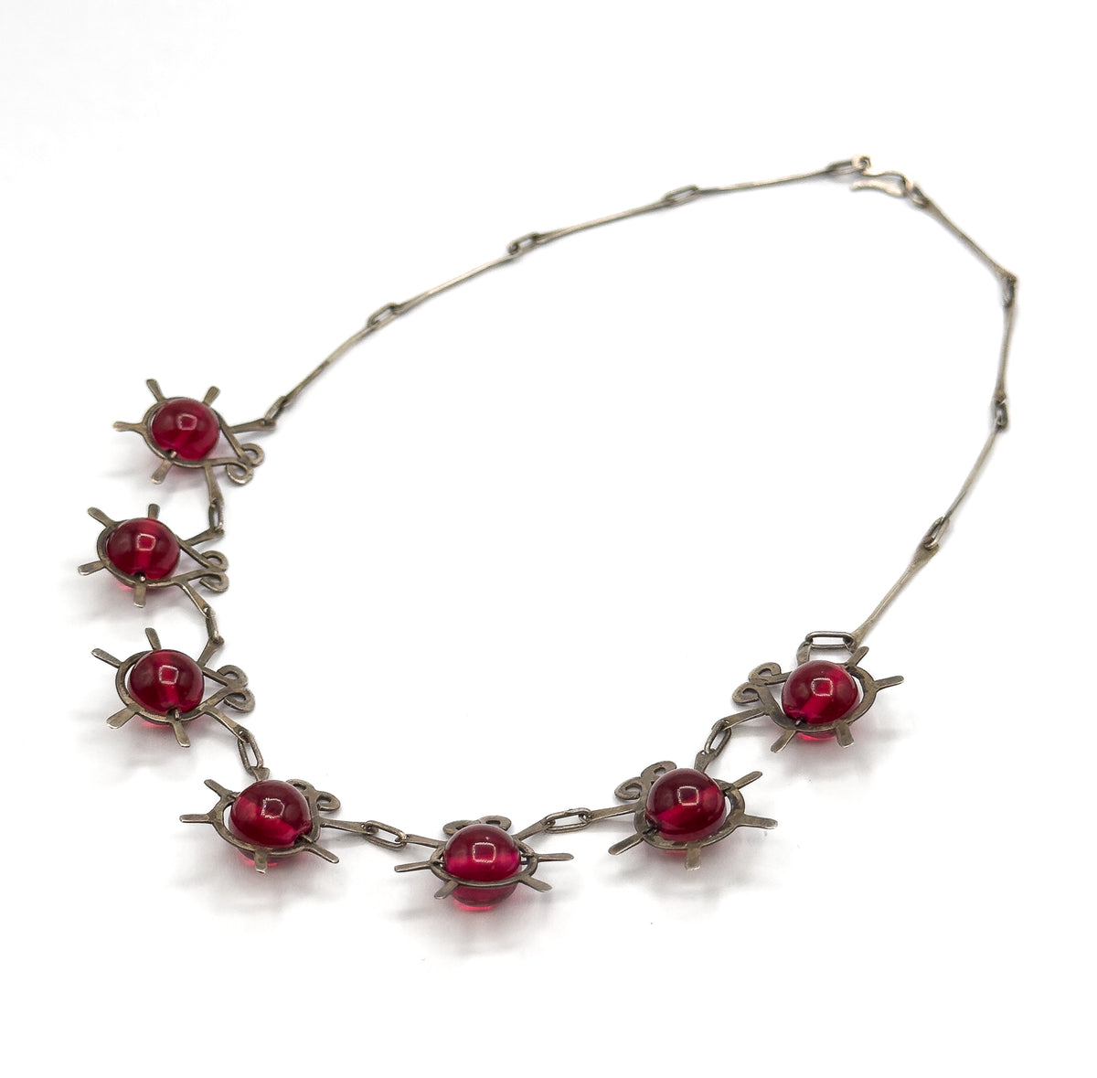 The necklace alone, chain looping up to its hook clasp at the top right, with all eight atomic insect stations and their cranberry glass beads spread along the lower curve.
