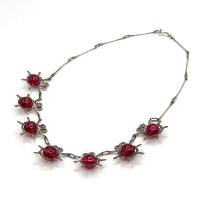 The necklace alone, chain looping up to its hook clasp at the top right, with all eight atomic insect stations and their cranberry glass beads spread along the lower curve.