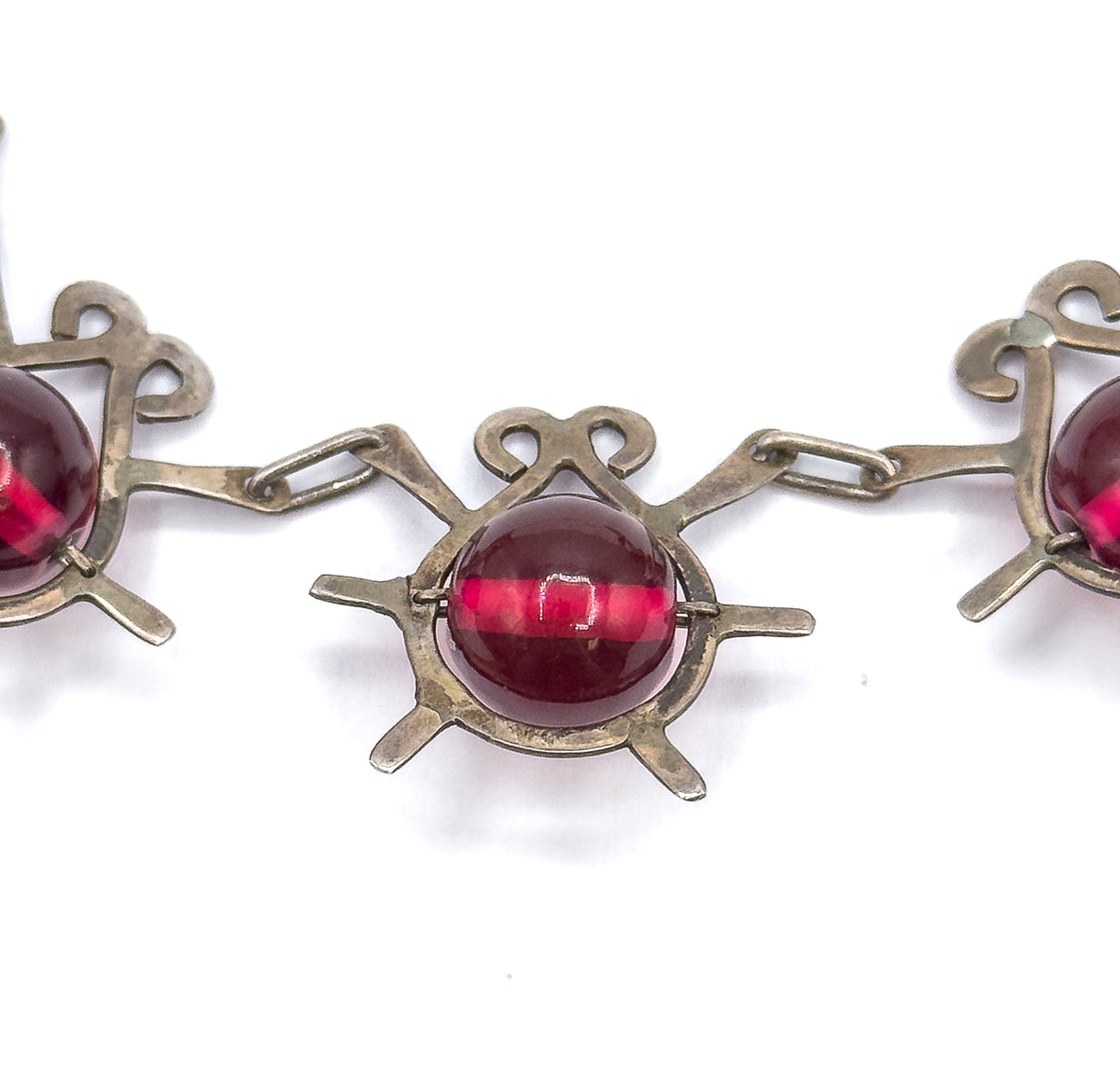 Tight macro on one atomic insect station, its curled antennae above and radiating sterling legs below, with slim prongs gripping the round cranberry glass bead between two elongated links.