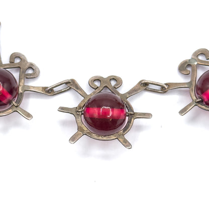 Tight macro on one atomic insect station, its curled antennae above and radiating sterling legs below, with slim prongs gripping the round cranberry glass bead between two elongated links.