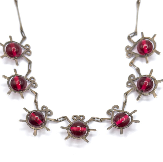 Seven atomic insect stations curve across the frame in the necklace's front section, their cranberry glass beads and radiating sterling legs joined by elongated links on white.