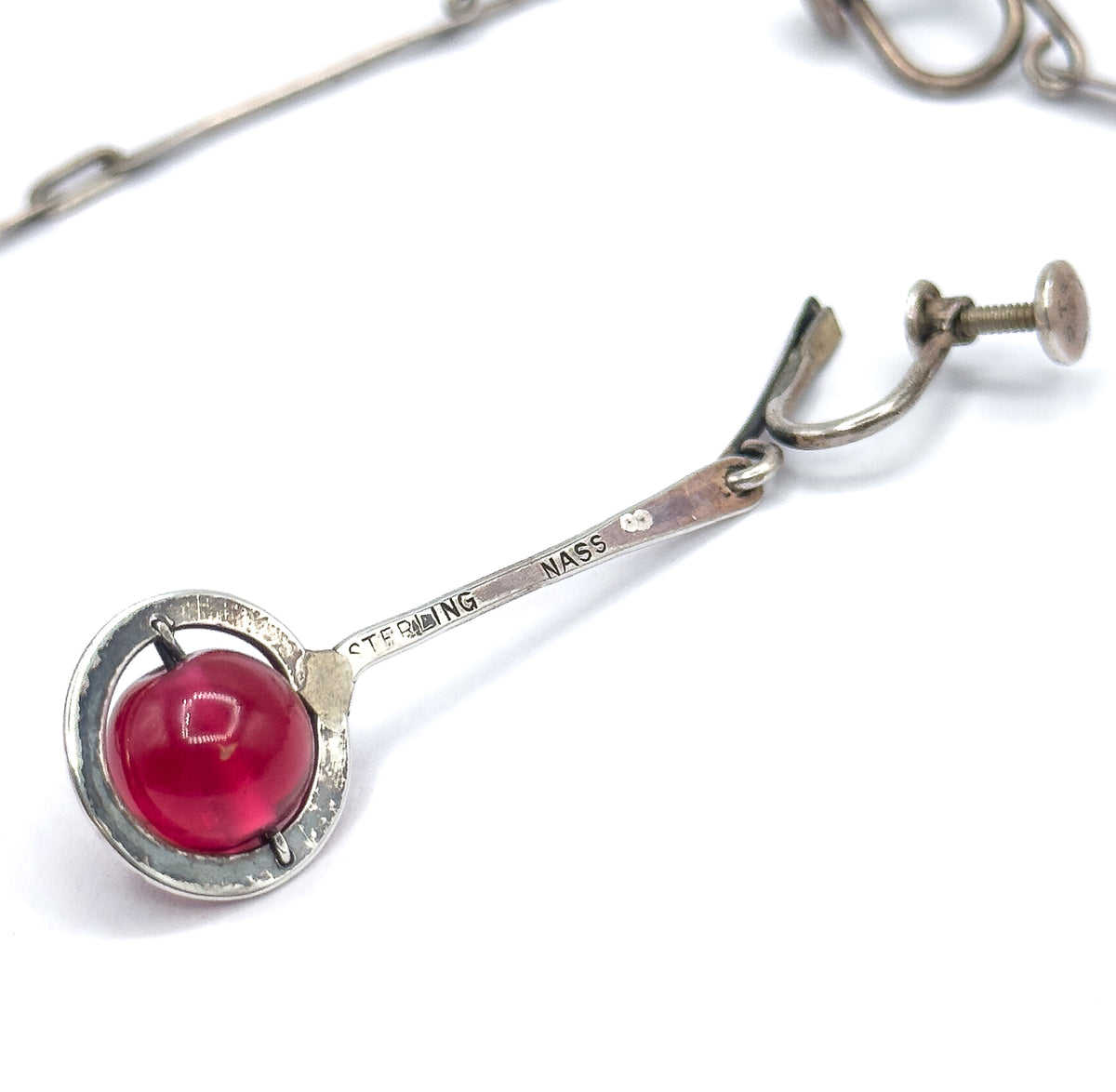 One earring lies diagonally with "STERLING" and "NASS" stamped along its flat sterling bar, ending in a circular frame where prongs hold the round cranberry glass bead, screwback above.