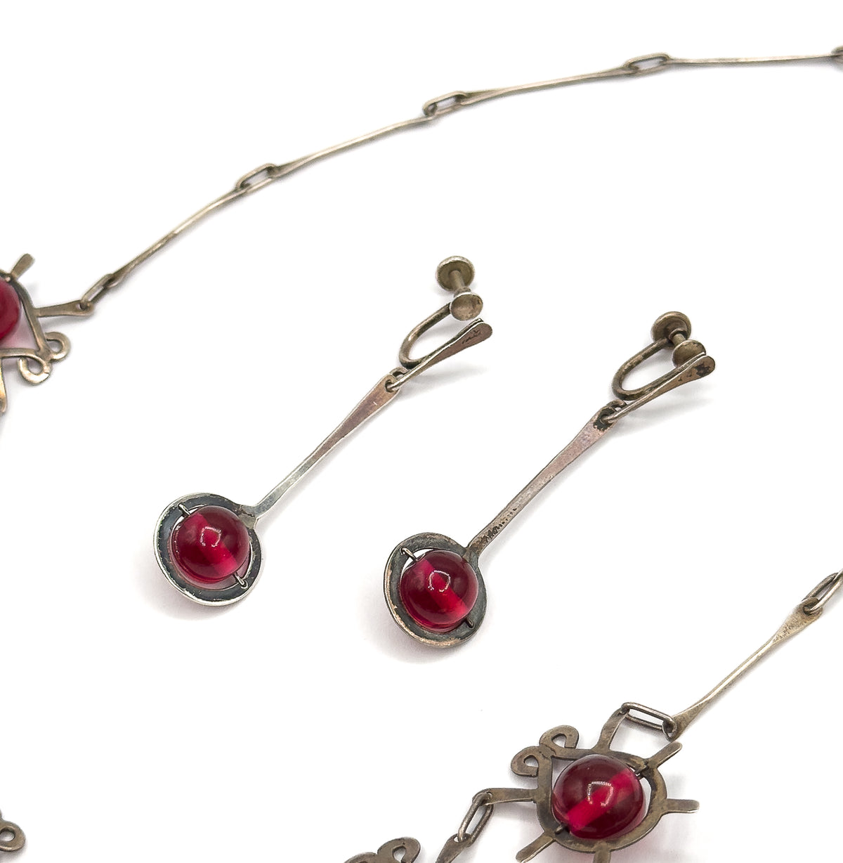 Both earrings lie side by side between the necklace's elongated sterling links, their long flat bars and cranberry glass drops seen alongside two atomic insect stations at the frame's edges.