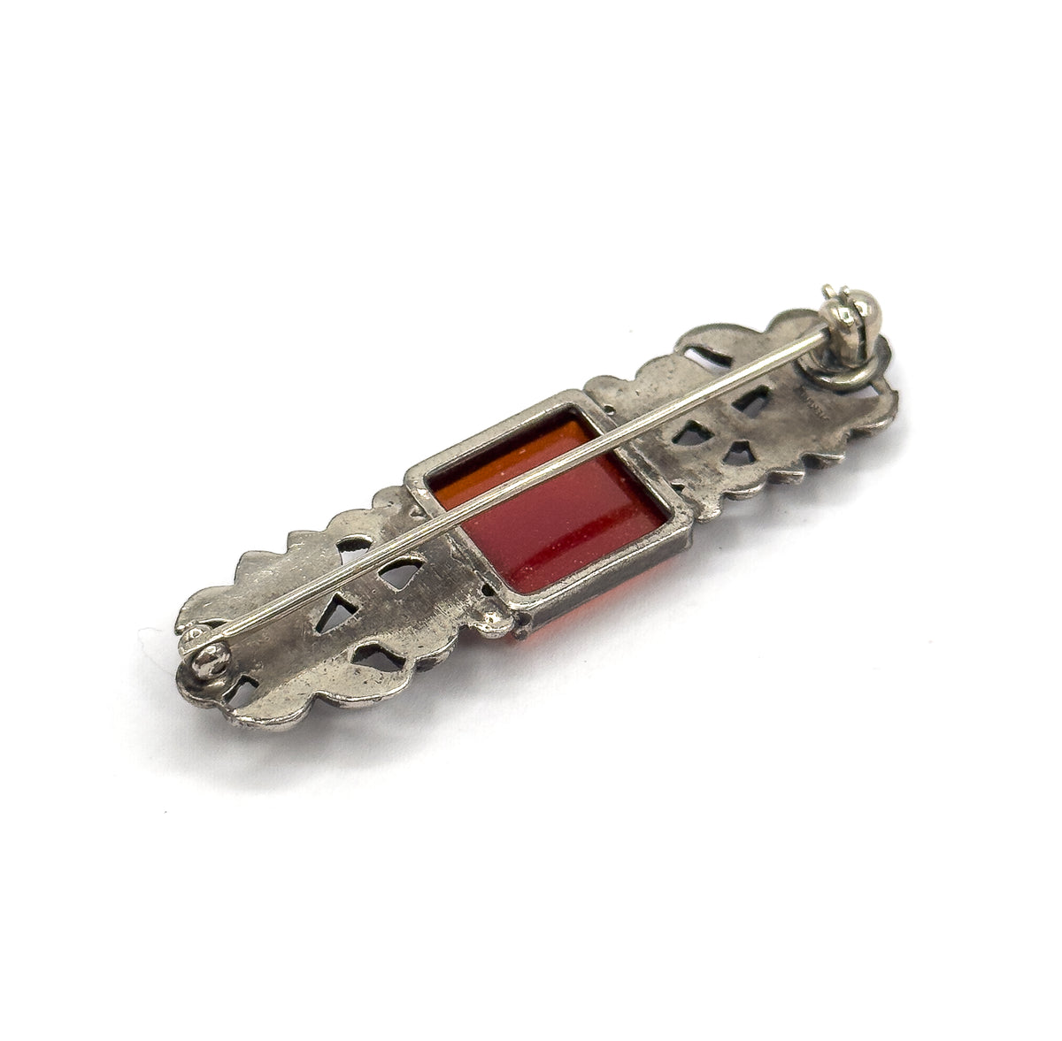 The reverse tilted lengthwise on white, the long pin stem running the full bar to its hinge at one end and the carnelian glowing orange-red through the open back of the bezel.