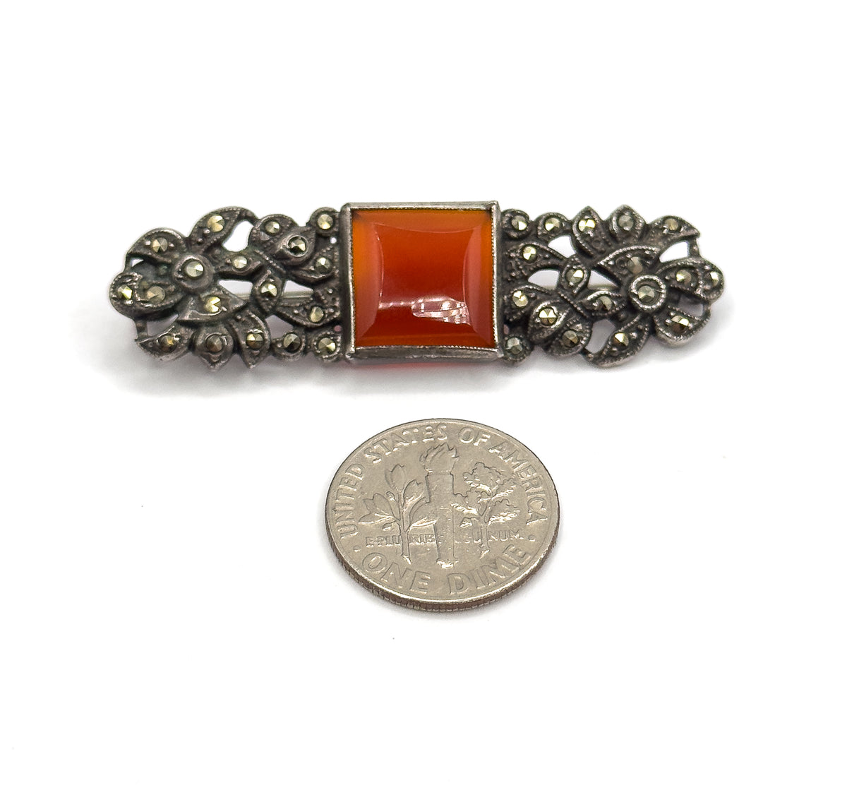 The brooch face up beside a dime for scale, the bezel-set square carnelian centred between marcasite-studded floral silver ends spanning about twice the coin's width.