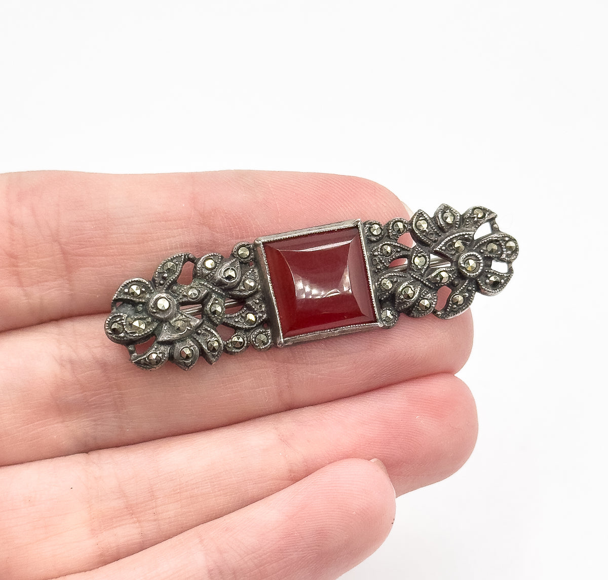 An Art Deco carnelian and marcasite sterling silver brooch resting across open fingers for scale, its bezel-set square stone flanked by darkened floral silverwork.