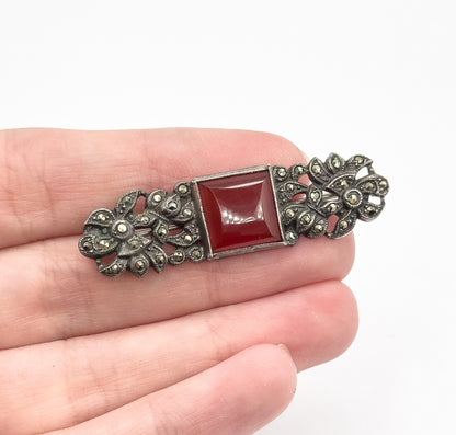 An Art Deco carnelian and marcasite sterling silver brooch resting across open fingers for scale, its bezel-set square stone flanked by darkened floral silverwork.