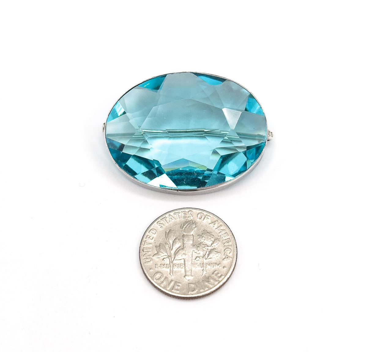 The brooch lies face up on white beside a dime, the coin roughly two-thirds the height of the oval paste stone, with the sterling silver bezel and pin catch just visible at the sides.