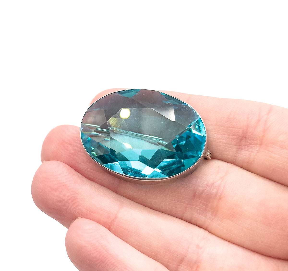 An Edwardian oval brooch with a large faceted topaz blue paste stone in a sterling silver bezel, resting on open fingers that show its size, C clasp visible at the side.