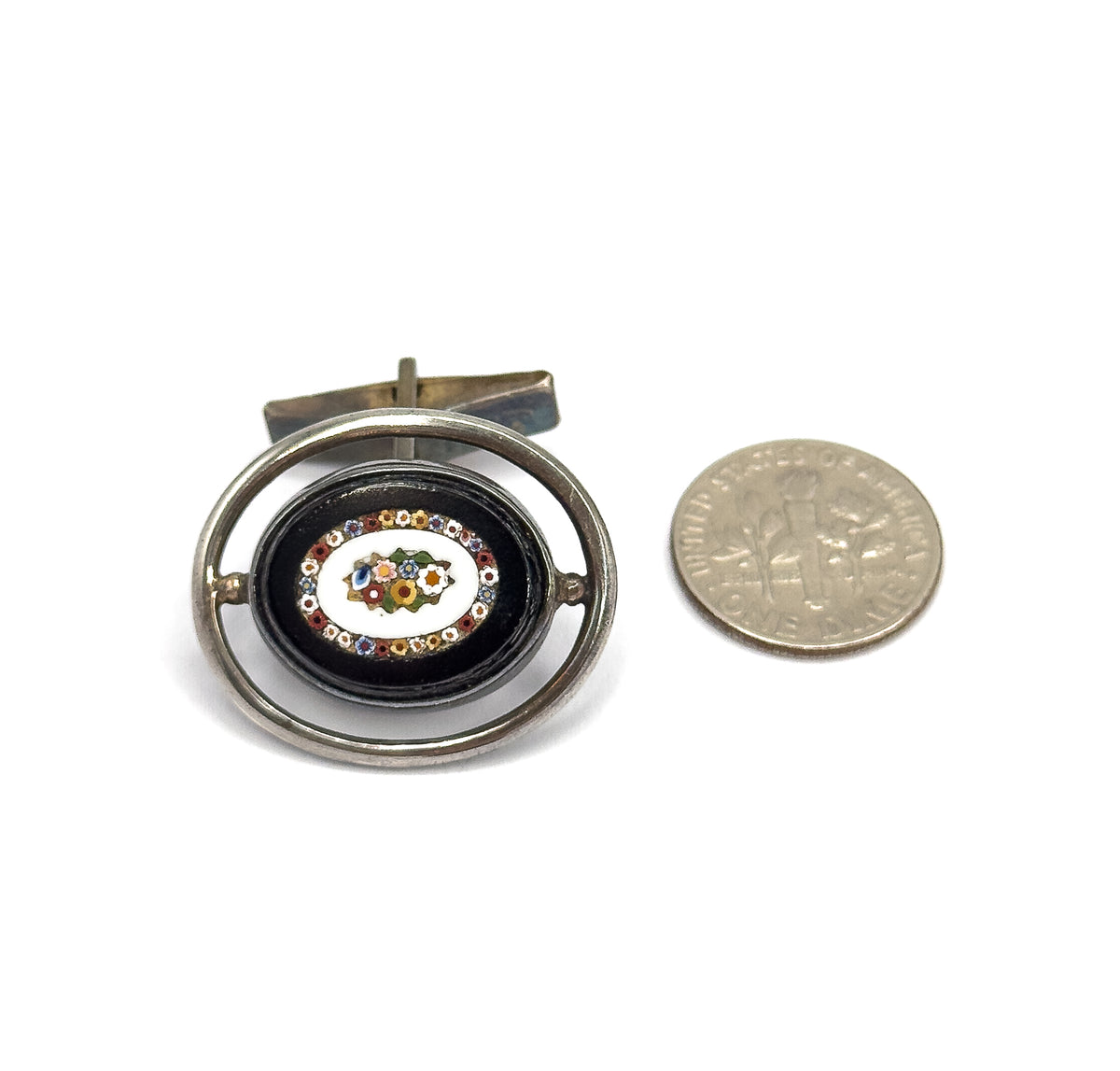 One cufflink lies face up beside a dime for scale, its oval mosaic of tiny glass flowers framed in black within a sterling silver hoop, toggle bar visible behind.