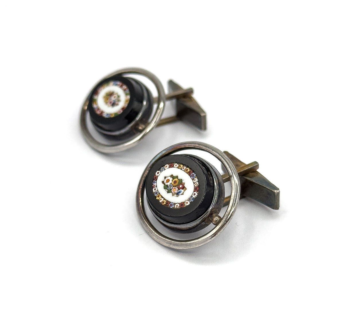 Front three-quarter view of both cufflinks, showing the multicoloured glass mosaic flower set in a pale ring at the centre of each dark oval face, encircled by a silver hoop.