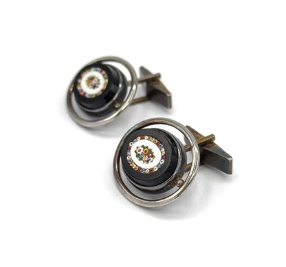 Front three-quarter view of both cufflinks, showing the multicoloured glass mosaic flower set in a pale ring at the centre of each dark oval face, encircled by a silver hoop.