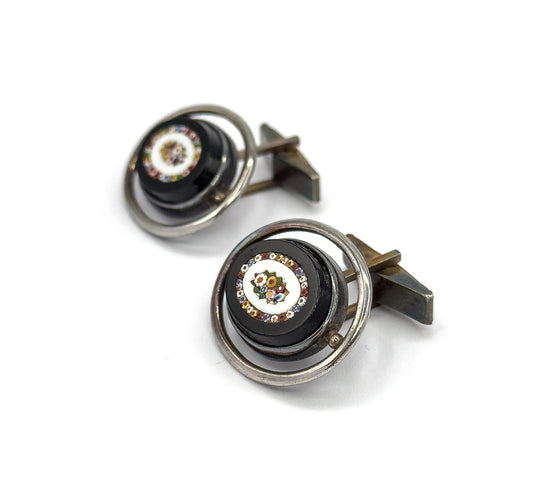 Front three-quarter view of both cufflinks, showing the multicoloured glass mosaic flower set in a pale ring at the centre of each dark oval face, encircled by a silver hoop.