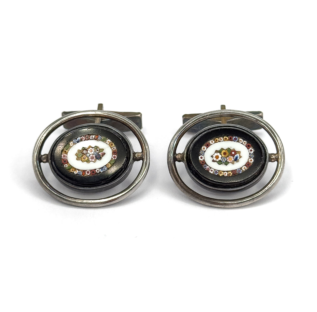 Straight-on front view of the pair side by side, each dark oval face showing its glass mosaic flower ringed by tiny multicoloured tesserae, with the toggle bars visible behind the silver hoops.