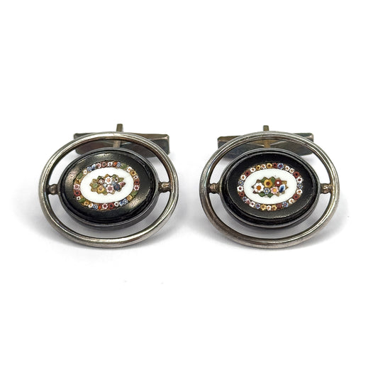 Straight-on front view of the pair side by side, each dark oval face showing its glass mosaic flower ringed by tiny multicoloured tesserae, with the toggle bars visible behind the silver hoops.