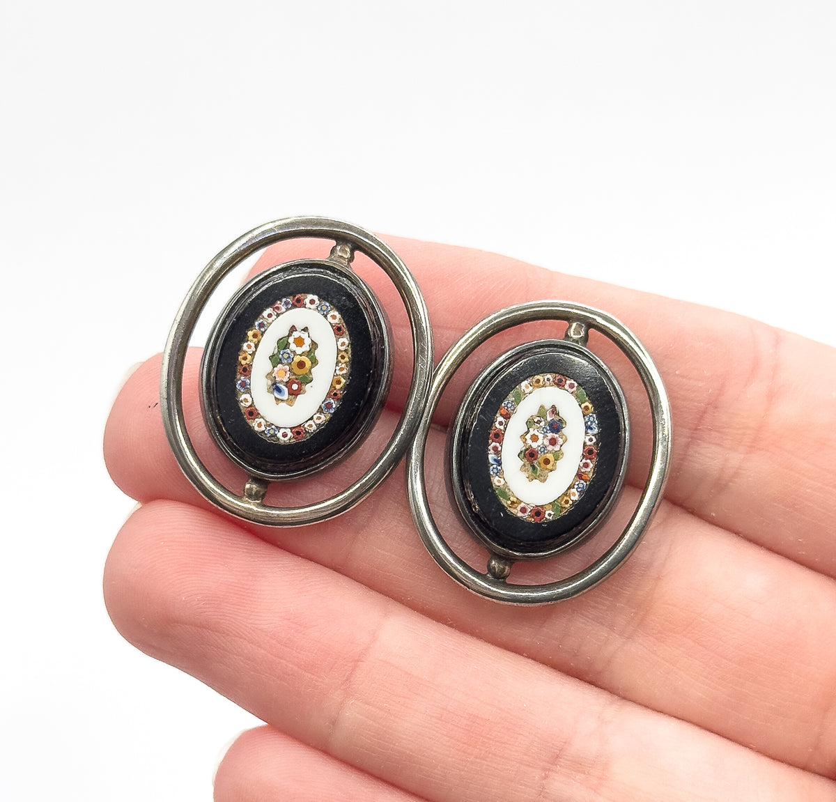 Sterling silver micro mosaic cufflinks, shown resting on fingertips for scale, each oval face ringed by a floating silver hoop and centred with a multicoloured glass flower design on white.