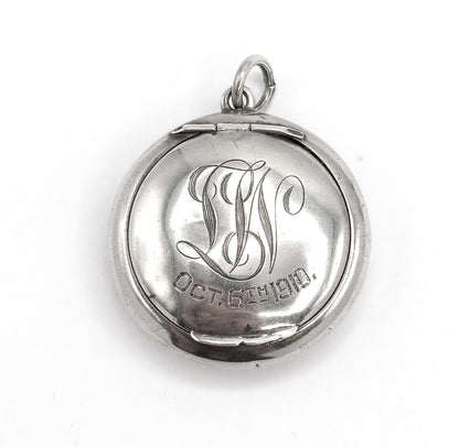 Straight-on view of the closed front filling the frame, the hinge and small catch tabs visible at top and bottom edges, script initials and engraved date centred beneath the round bail ring.