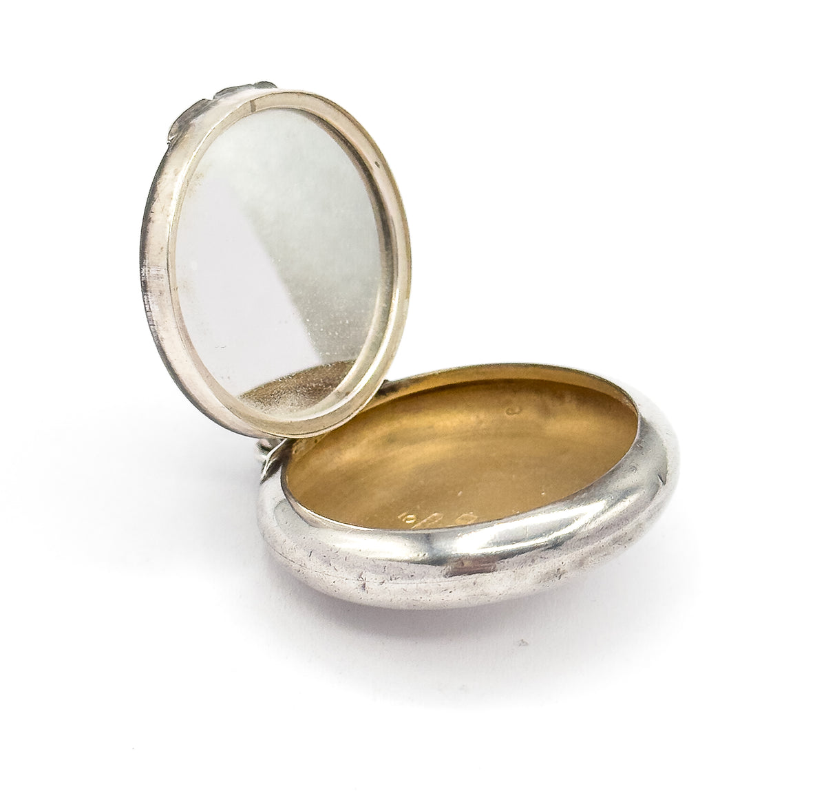 The locket shown open against white, the hinged lid lifted to reveal the mirror inside, the deep rounded base showing a warm-toned interior and small marks stamped near its rim.