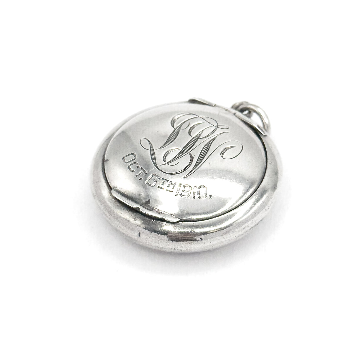 Angled from the opposite side, the engraved lid sits within the wider rim of the base, the seam running along the lower edge and the bail ring at the upper right, script initials and date upright.
