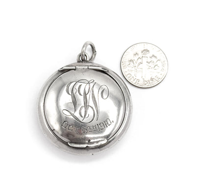 The engraved front shown flat beside a dime for scale, the locket roughly three times the coin's width, its script initials and date centred below the bail ring.