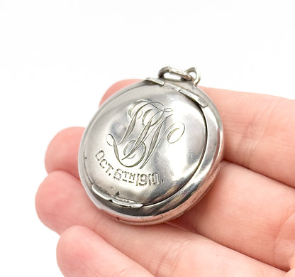 Edwardian round sterling silver locket, shown resting on fingertips for scale, engraved script initials and a date across the polished front, with a ring-shaped bail at the top.