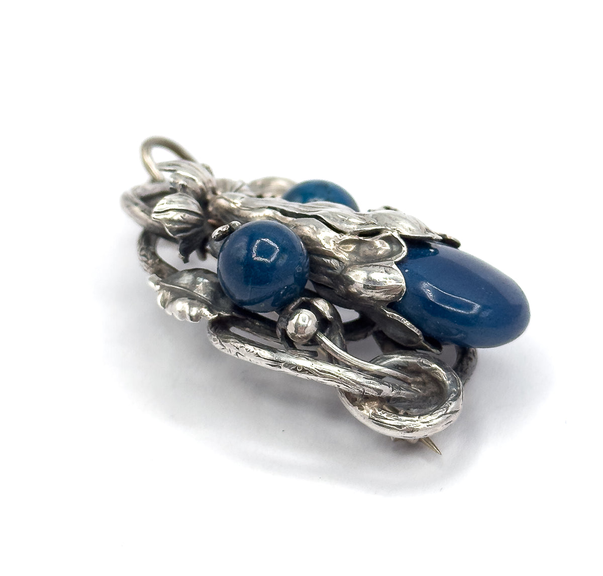 Angled three-quarter view from the pin side, the long hooked catch and pin stem curving across the lower edge while the oval sodalite cabochon and two round beads sit raised behind.