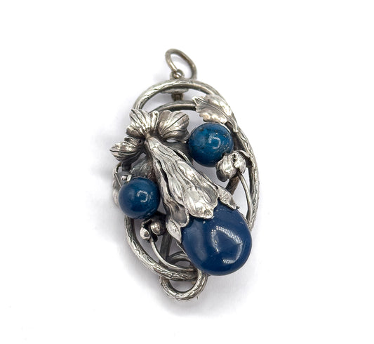 Upright front view against white, the whole oval openwork frame visible with its curling vine tendrils and small buds encircling the three deep blue stones beneath the bail loop.
