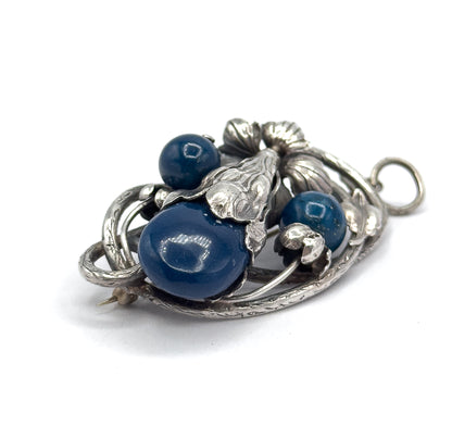 Close tilted view from below the left edge, the pin stem tip just visible under the frame while light catches the domed face of the large sodalite cabochon and the bail ring sits far right.