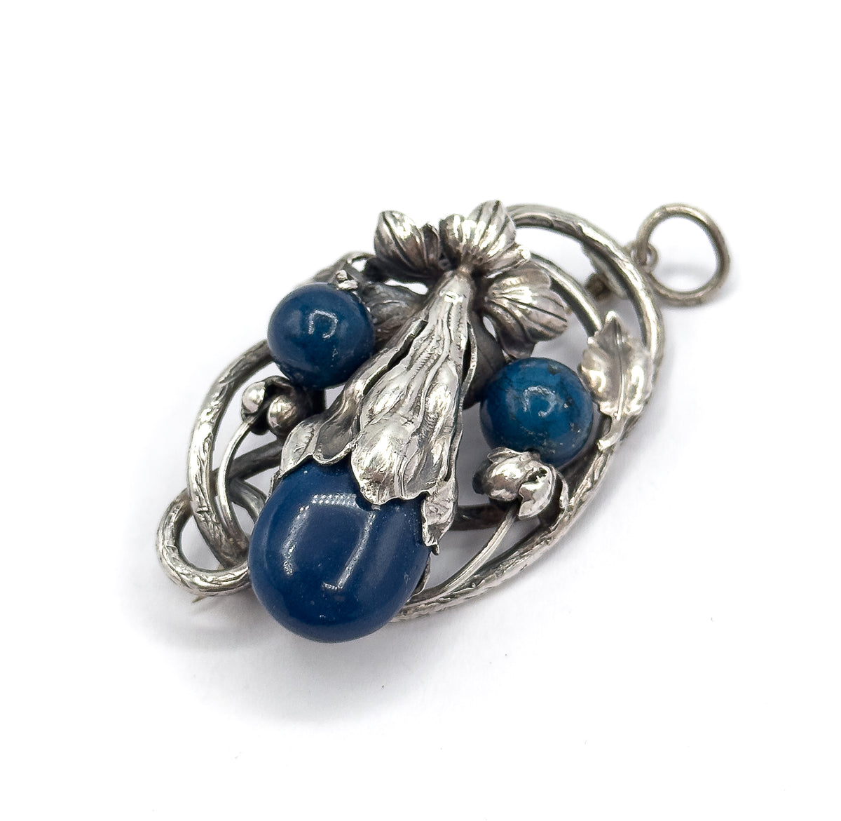 Tilted diagonal view with the bail ring at upper right, showing the sculpted lily flower and long ribbed leaf standing proud at the centre between the two round beads and the large oval cabochon.