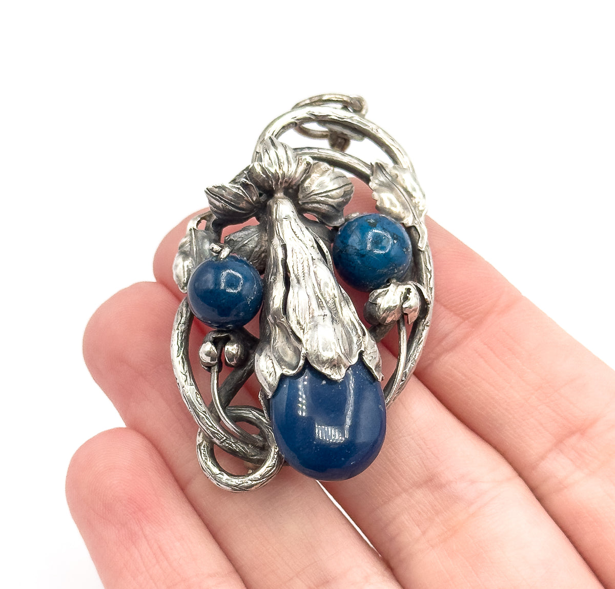 Art Nouveau sterling silver flower pendant, a large deep blue sodalite cabochon and two round blue beads within a curving floral vine frame, shown resting on open fingers for scale.