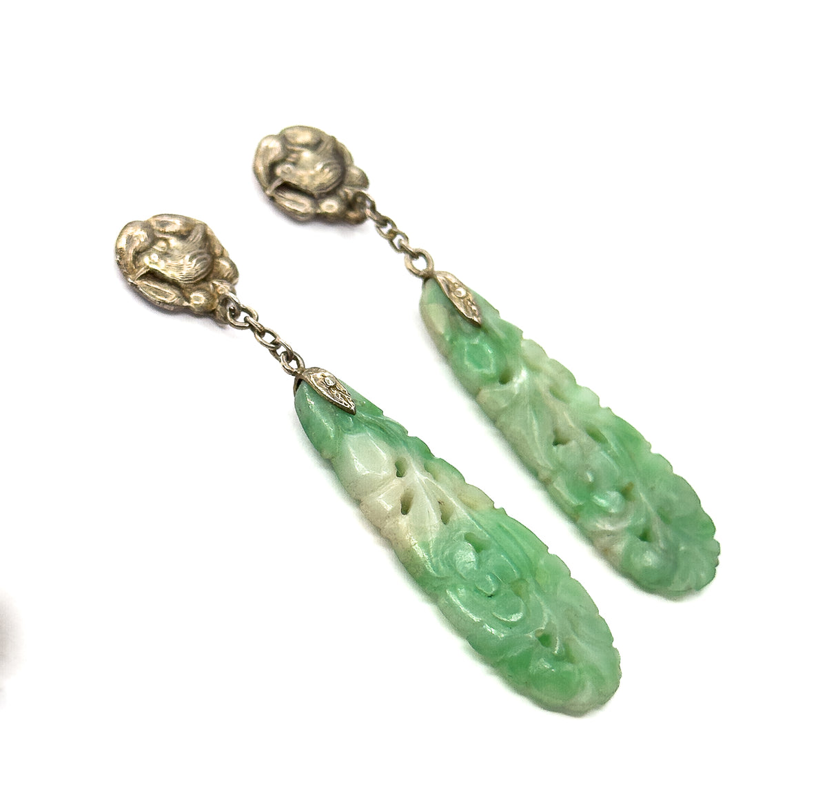 Both earrings on white angled diagonally with the drops separated, showing the long carved jade faces in profile-lit relief and the chain stretched taut between each bird top and metal cap.