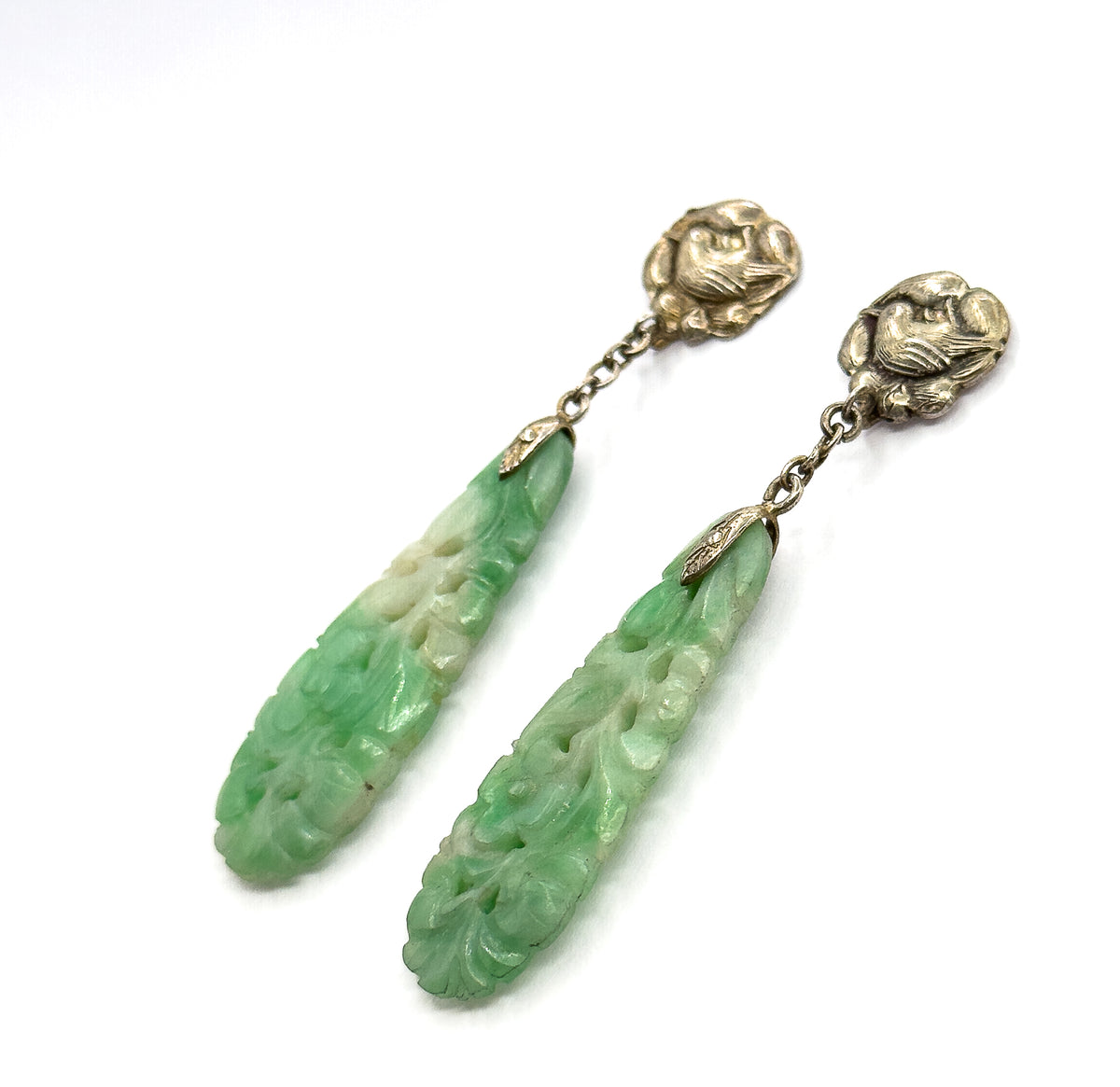Overhead diagonal view of the pair lying parallel across white, both bird-motif tops facing up at the top right and the tapering carved jade drops angling down to the lower left.