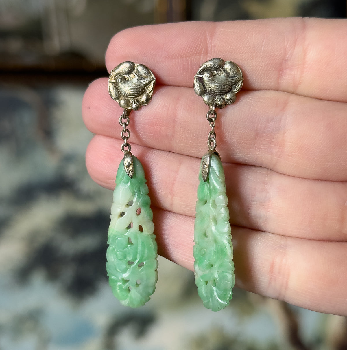 Art Deco carved jade drop earrings in sterling silver, shown resting across the fingers of an open hand, each embossed bird top linking by chain to a long minty green carved drop.