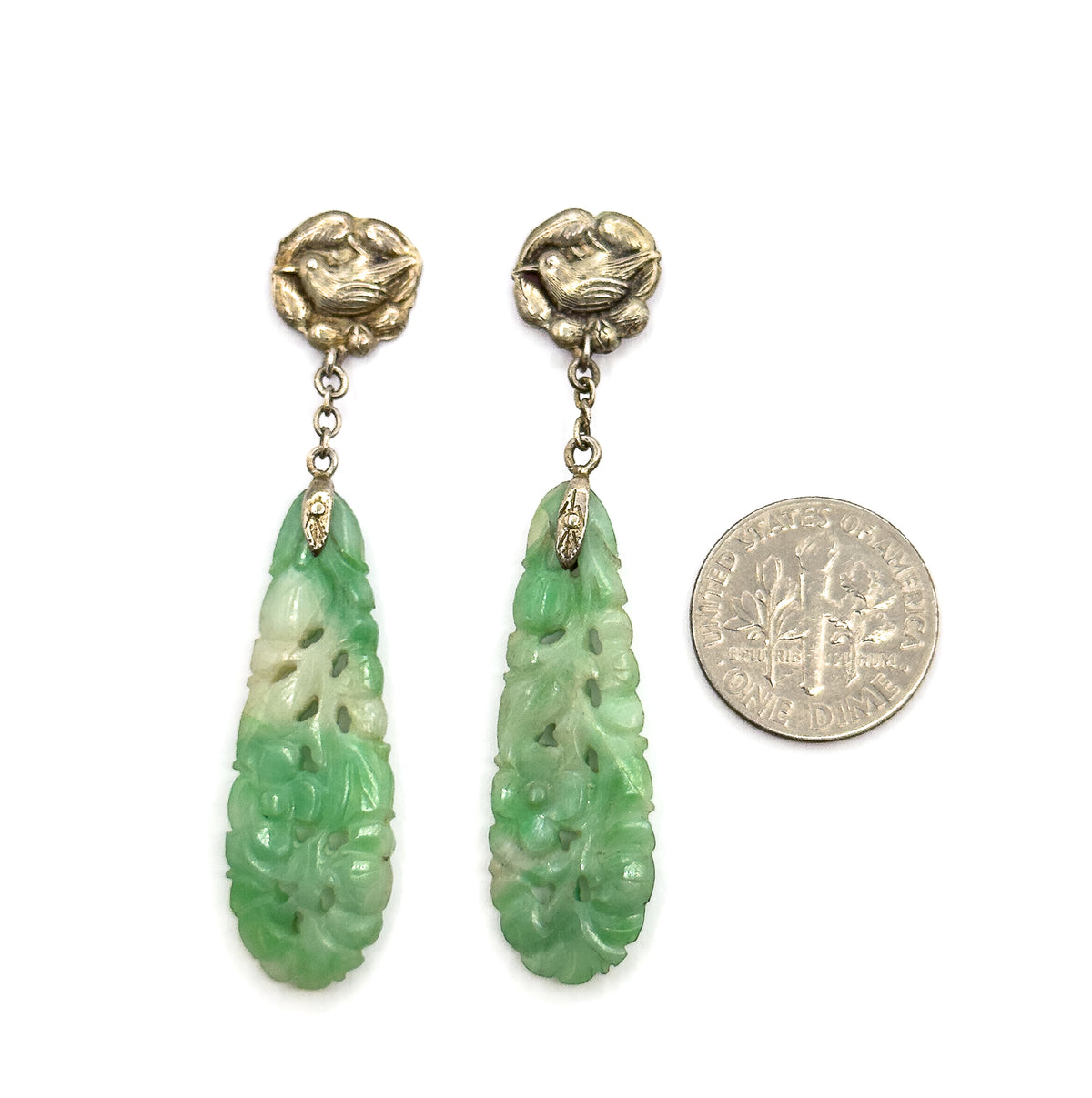 Both earrings laid flat beside a dime for scale, the coin roughly matching the length of one carved jade drop below its chain and bird top.