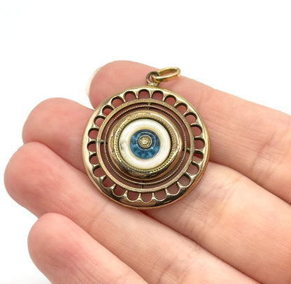 Art Deco mother of pearl and blue glass pendant in gold fill, shown resting on the fingers of a hand for scale, its scalloped round target design and bail facing the camera.