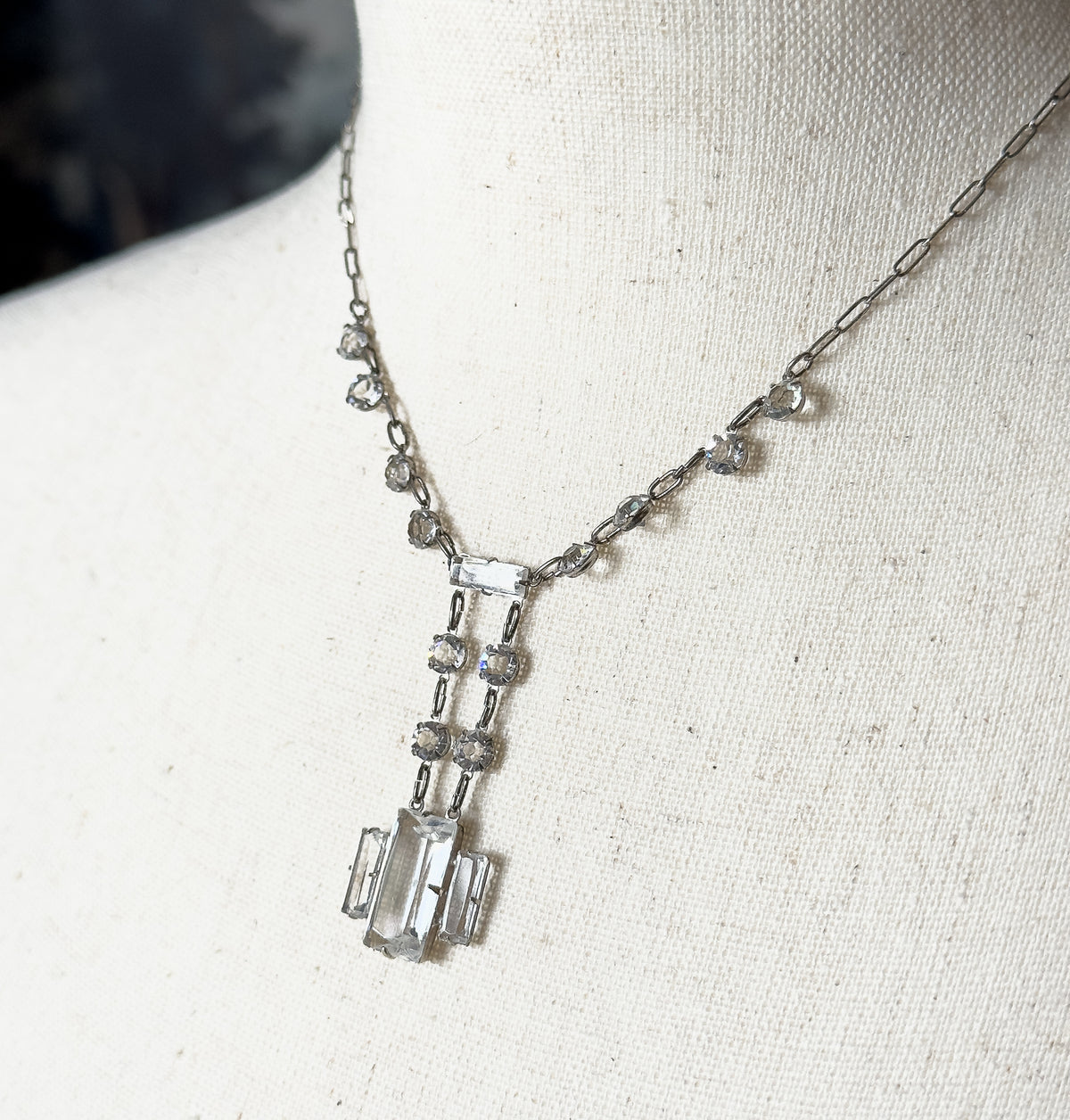 An Art Deco colorless cut glass necklace in sterling silver and base metal, shown at the neck of a cream fabric bust form, its rectangular centre stones hanging in a linear geometric drop.