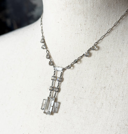 An Art Deco colorless cut glass necklace in sterling silver and base metal, shown at the neck of a cream fabric bust form, its rectangular centre stones hanging in a linear geometric drop.