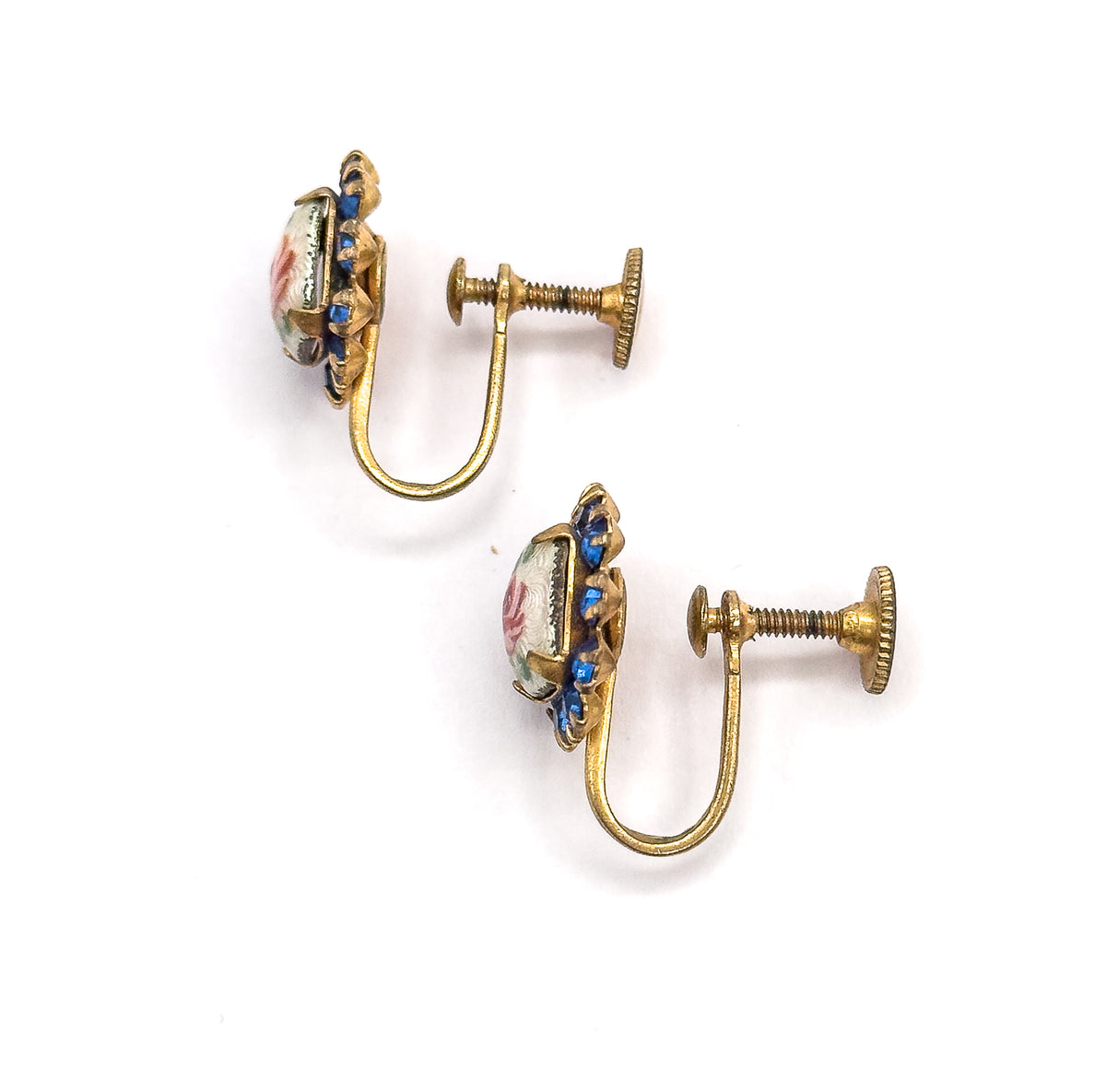 Angled side view of the pair, showing the shallow depth of each enamel rose face, the cobalt blue glass stones edging the frame, and the screw discs extended along their threads.