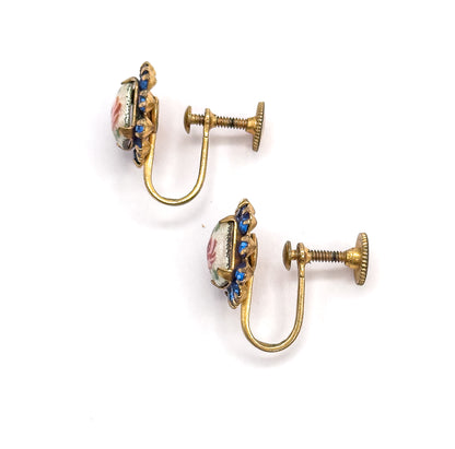 Angled side view of the pair, showing the shallow depth of each enamel rose face, the cobalt blue glass stones edging the frame, and the screw discs extended along their threads.