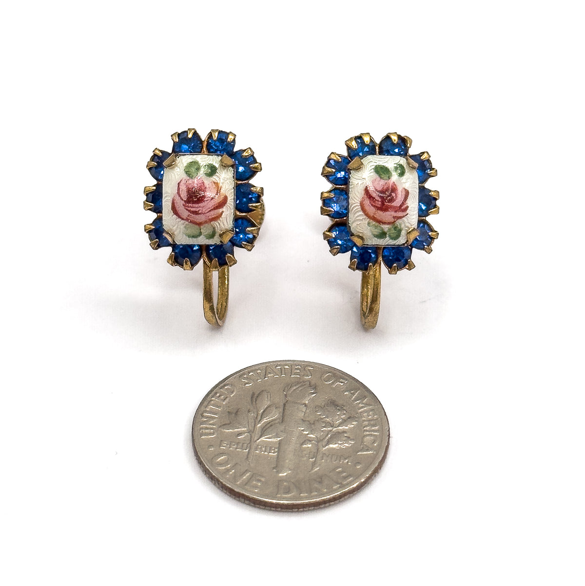 The pair face-up beside a dime for scale, each hand-painted enamel rose and its cobalt blue glass halo sitting a little wider than half the coin, with the curved screw fittings below.