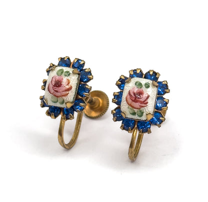 Close three-quarter view of the pair standing upright, the hand-painted enamel roses tilted toward the camera within their prong-set cobalt blue glass halos, curved screw wires below.