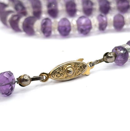 Close macro of the oval base metal clasp with raised scrolling decoration, linked at each end to small round metal beads and amethyst rondelles, with the strand blurred behind.