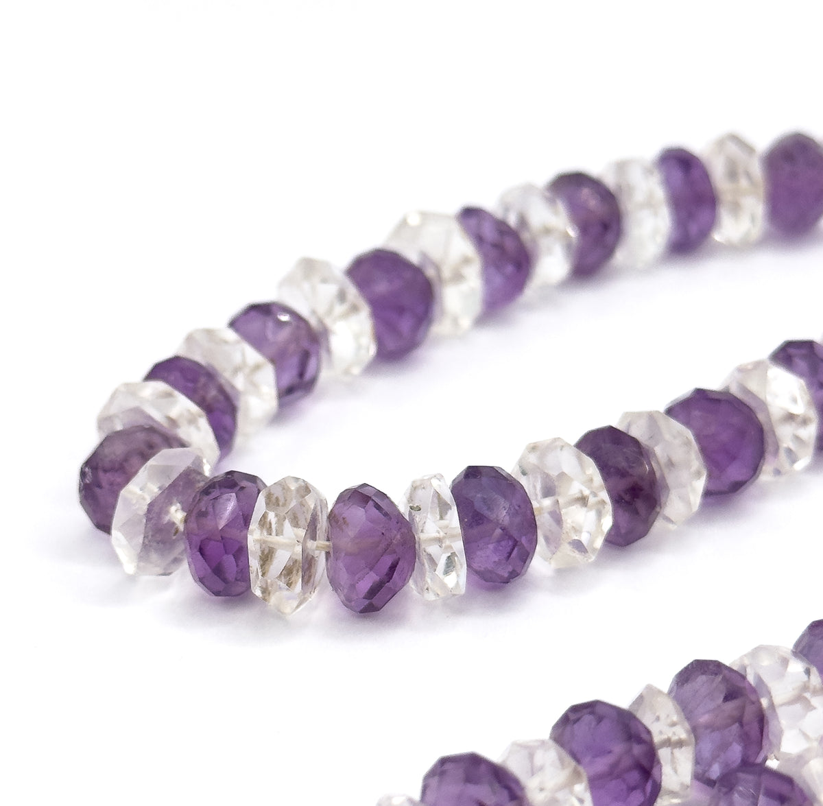 Two sweeping loops of the strand curve across a white ground, the nearest amethyst and rock crystal rondelles in sharp focus and noticeably broader than those receding behind.