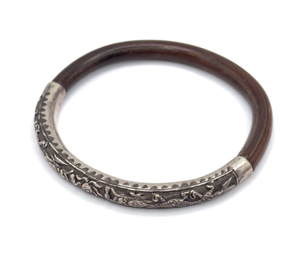 A single bangle lies flat on white, the deep brown wood band sweeping round to a sterling silver section embossed with raised figures and a toothed engraved border.