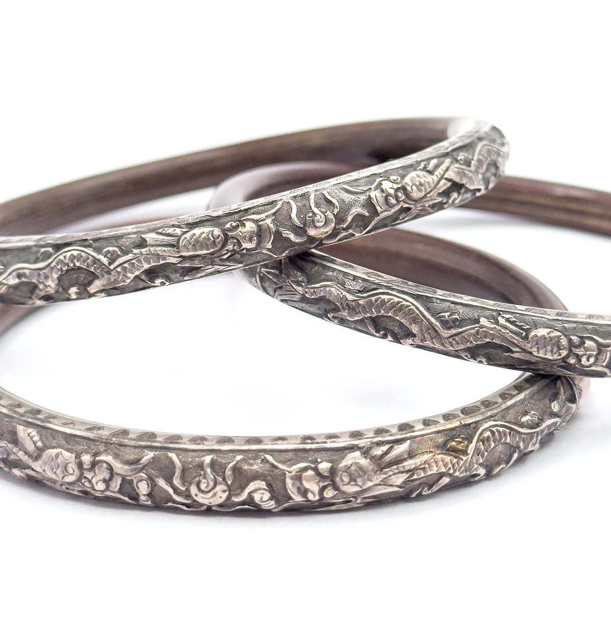 Tight crop across the three stacked bangles, the raised embossed figures and scrolling clouds standing proud of the silver with patina darkening the recesses and brown wood curving behind.