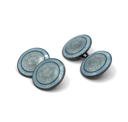 Both cufflinks stand angled on a white background, each pair of discs linked by its toggle bar, the dusty blue rims and central rosette pattern catching the light in three-quarter view.