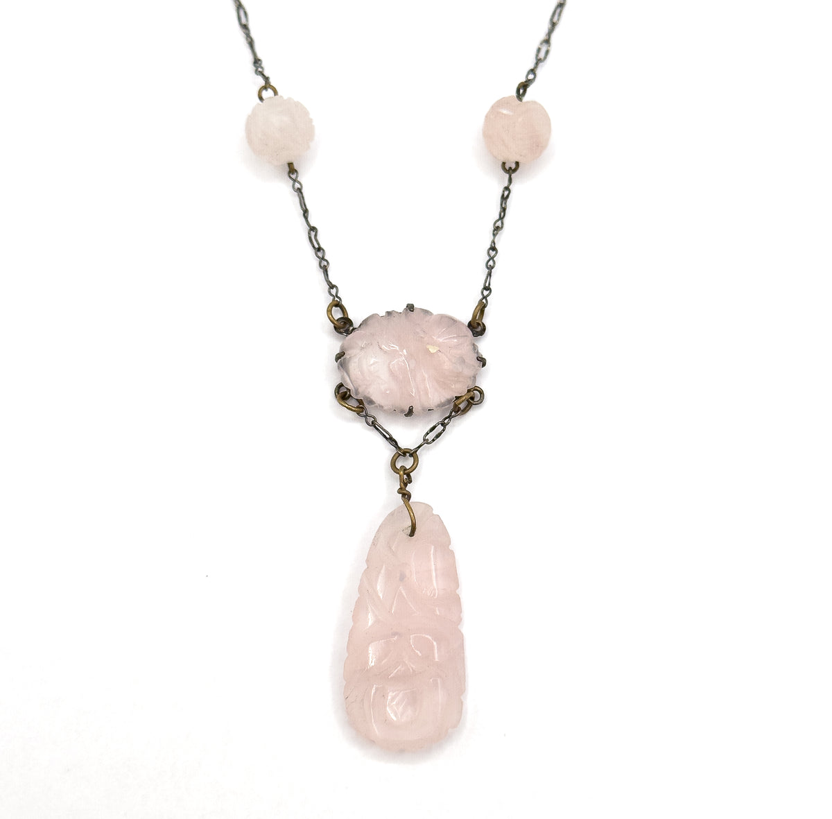 Straight-on symmetrical view of the necklace front on white, both round carved rose quartz beads balanced either side of the pronged oval centre station with the long carved drop hanging centred below.