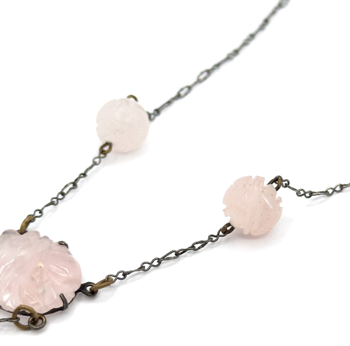 Macro detail of the darkened chain links running between two hand-carved rose quartz beads, with the edge of the pronged oval centre stone entering at lower left.